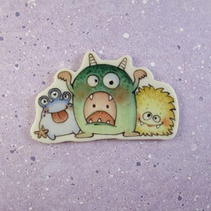 May include: Three cartoon monsters with different features and colors. The monster on the left has four eyes and a blue body, the middle monster is green with a wide open mouth and two horns, and the monster on the right is yellow and fluffy.
