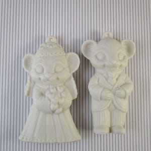 May include: A pair of white ceramic mouse figurines, one dressed as a bride and the other as a groom. The bride is holding a bouquet of flowers and the groom is holding a small book.