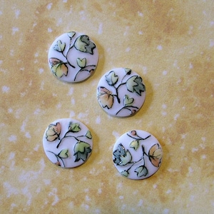 May include: Four white ceramic buttons with a floral design. The buttons feature a white background with green and orange leaves and stems.