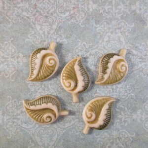 May include: Five ceramic leaf-shaped beads. Each bead features a cream-colored base with olive green and white accents. The leaves have detailed veining and a curled design. The beads are arranged on a light blue patterned surface.