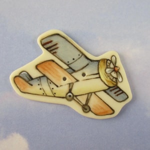 May include: A whimsical, hand-painted airplane pin. The biplane features a light yellow body, gray wings, and orange accents. The propeller is yellow with a red center. The pin is set against a light blue background.