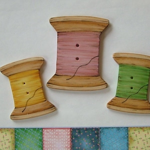 May include: Three wooden spool buttons with painted thread. The spools are painted in shades of pink, yellow, and green. The buttons are approximately 2 inches in diameter.
