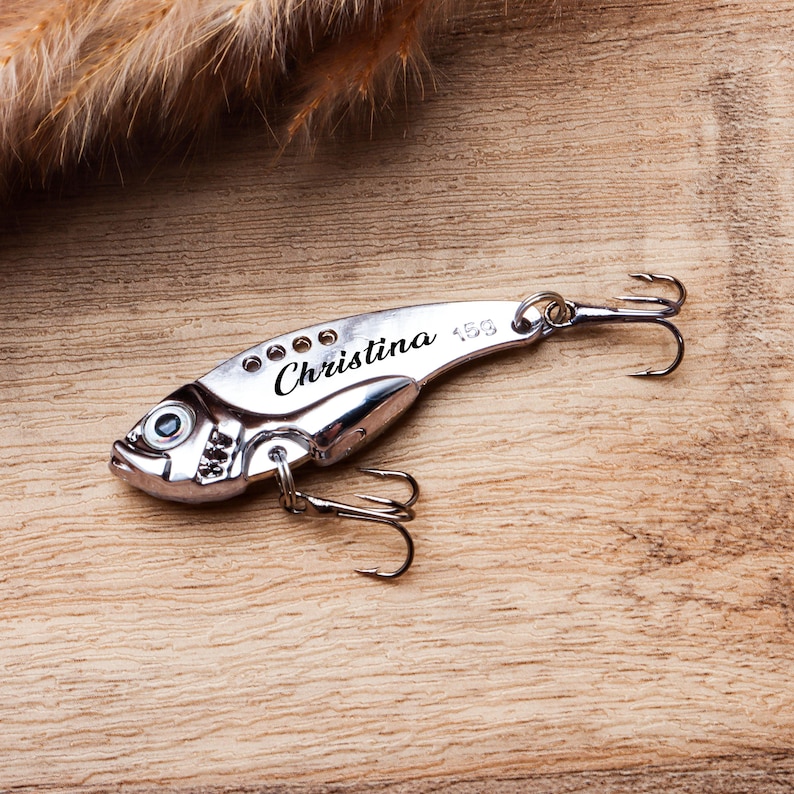 Pode incluir: Uma isca de pesca prateada com o nome "Christina" gravado na lateral. A isca tem formato de peixe com um anzol e est&aacute; sobre um fundo de madeira. A isca foi projetada para atrair peixes.