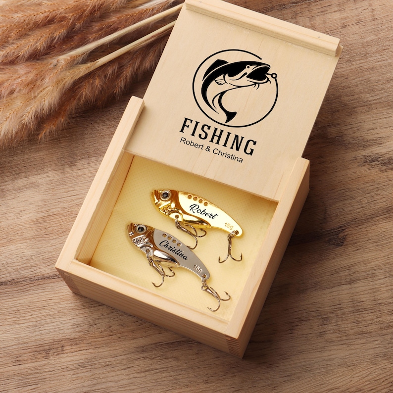 Pode incluir: Caixa de presente de madeira com tampa articulada, com um logotipo de peixe em preto e branco e a palavra "FISHING". No interior, s&atilde;o apresentadas duas amostras de pesca personalizadas. Uma &eacute; dourada, a outra prateada, cada uma gravada com um nome.
