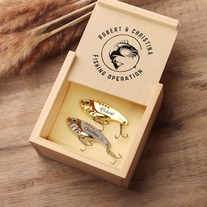 Pode incluir: Caixa de madeira com tampa articulada, contendo duas iscas de pesca. A tampa &eacute; gravada com "Robert & Christina Fishing Operation" e um gr&aacute;fico de peixe. Uma isca &eacute; dourada, a outra prateada, ambas com anz&oacute;is e nomes personalizados.