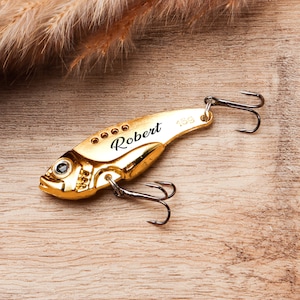 Pode incluir: Uma isca de pesca dourada em forma de peixe, com o nome "Robert" gravado. A isca tem um anzol na parte superior e inferior, e o n&uacute;mero "15g" est&aacute; impresso na isca. A isca est&aacute; sobre uma superf&iacute;cie de madeira.
