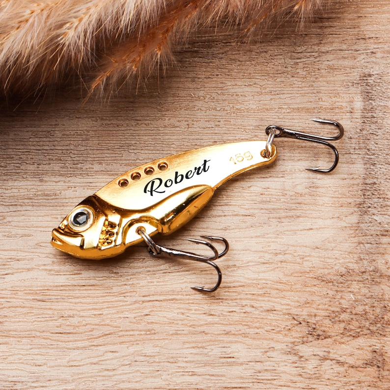 Pode incluir: Uma isca de pesca dourada em forma de peixe, com o nome "Robert" gravado. A isca tem um anzol na parte superior e inferior, e o n&uacute;mero "15g" est&aacute; impresso na isca. A isca est&aacute; sobre uma superf&iacute;cie de madeira.
