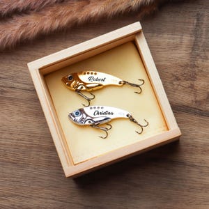 Pode incluir: Uma caixa de madeira cont&eacute;m duas amostras de pesca personalizadas. Uma &eacute; dourada com o nome "Robert" e a outra prateada com o nome "Christina". Ambas as amostras t&ecirc;m anz&oacute;is triplos e s&atilde;o exibidas sobre um fundo amarelo.