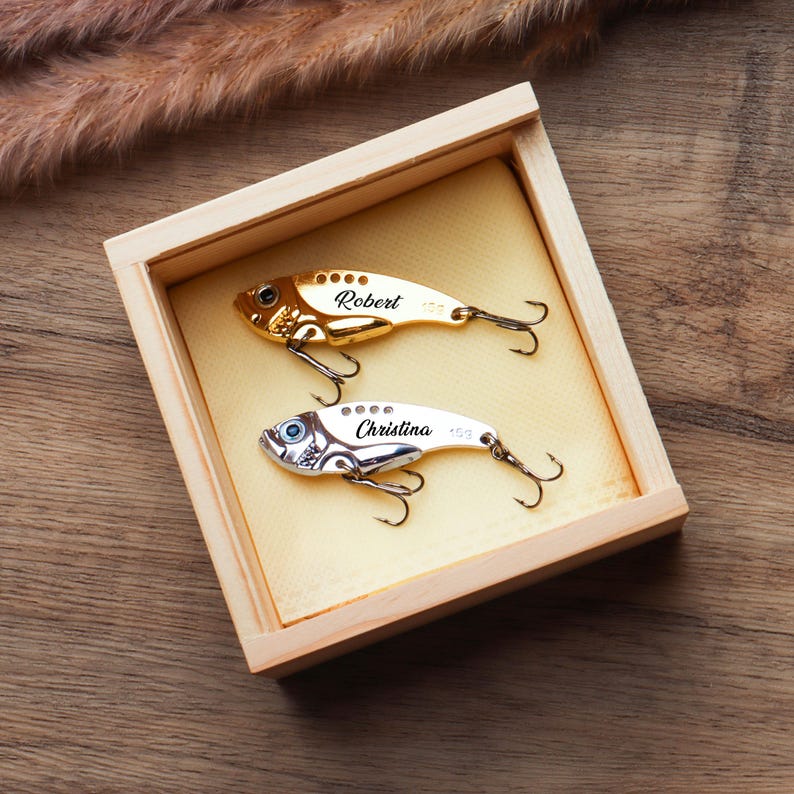 Pode incluir: Uma caixa de madeira cont&eacute;m duas amostras de pesca personalizadas. Uma &eacute; dourada com o nome "Robert" e a outra prateada com o nome "Christina". Ambas as amostras t&ecirc;m anz&oacute;is triplos e s&atilde;o exibidas sobre um fundo amarelo.