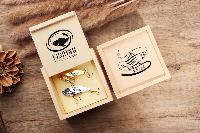 Pode incluir: Caixa de presente de madeira com tema de pesca com duas iscas. A tampa da caixa apresenta um gr&aacute;fico de peixe preto e a palavra "FISHING". As iscas s&atilde;o douradas e prateadas com os nomes "Robert" e "Christina".