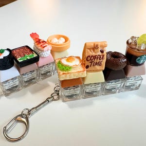 May include: Two rows of miniature food-themed keychains featuring sushi, shrimp nigiri, dim sum, toast with a fried egg, a coffee bag labelled COFFEE TIME, a chocolate doughnut, and a coffee cup with a green logo, all mounted on clear plastic keycaps.