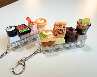 May include: Two rows of miniature food-themed keychains featuring sushi, shrimp nigiri, dim sum, toast with a fried egg, a coffee bag labeled COFFEE TIME, a chocolate donut, and a coffee cup with a green logo, all mounted on clear plastic keycaps.