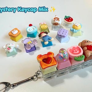 May include: A collection of colourful, miniature decorative keycaps featuring various shapes including animals, food items, and flowers. Some keycaps are attached to a silver metal keychain ring. The text Mystery Keycap Mix is visible.