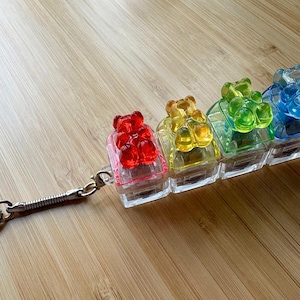 May include: A keyring featuring four translucent, square-shaped blocks, each topped with a small, colourful gummy bear figure in red, yellow, green, and blue. The keyring includes a silver-toned metal ring and a spring-like connector attachment.