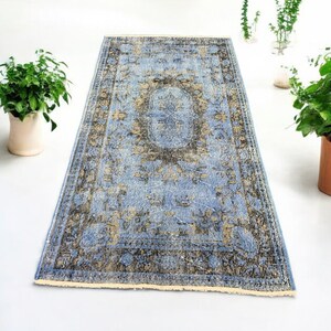 Puede incluir: Una alfombra rectangular de aspecto envejecido, con un campo azul claro y complejos diseños florales en tonos marrón y dorado. La alfombra tiene un diseño de medallón central y un borde oscuro. Colocada sobre una superficie blanca.