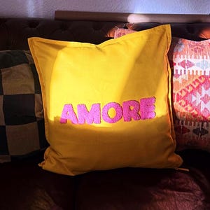 May include: Three decorative throw pillows on a brown leather couch. The center pillow is yellow with the word "AMORE" in pink. Other pillows feature a checkered pattern and a geometric design.