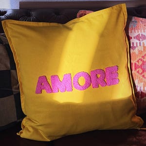 Könnte beinhalten: Ein senfgelbes Zierkissen mit dem Wort "AMORE" in Pink gestickt. Das quadratische Kissen liegt auf einem braunen Ledersofa, mit anderen gemusterten Kissen im Hintergrund.