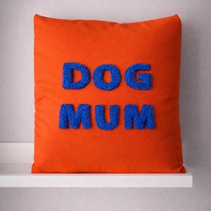 Könnte beinhalten: Ein orangefarbenes quadratisches Kissen mit den Worten "DOG MUM" in blauen, strukturierten Buchstaben. Das Kissen wird auf einem weißen Regal vor einem neutralen Hintergrund präsentiert. Ein dekorativer Artikel für Hundeliebhaber.