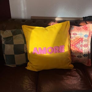 May include: Three decorative throw pillows on a brown leather couch. The center pillow is yellow with the word "AMORE" in pink. A checkered pillow and a patterned pillow are also visible.
