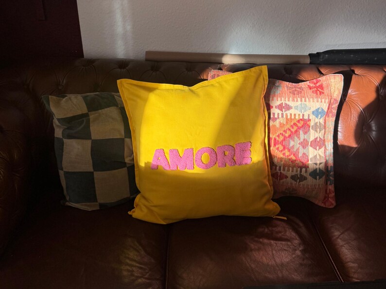 May include: Three decorative throw pillows on a brown leather couch. The center pillow is yellow with the word "AMORE" in pink. A checkered pillow and a patterned pillow are also visible.