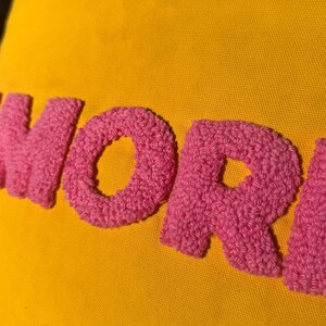 May include: Close-up of a yellow fabric with the word "MORE" embroidered in pink, textured yarn. The letters are large and stand out against the bright background. The image is well-lit, highlighting the texture.