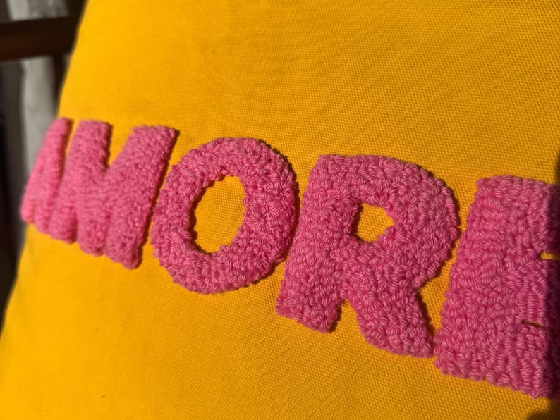May include: Close-up of a yellow fabric with the word "MORE" embroidered in pink, textured yarn. The letters are large and stand out against the bright background. The image is well-lit, highlighting the texture.