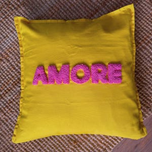 May include: A square yellow pillow with the word "AMORE" embroidered in pink, placed on a textured brown surface. The pillow has a simple design, with a solid yellow background and the word "AMORE" in a bold, textured font.