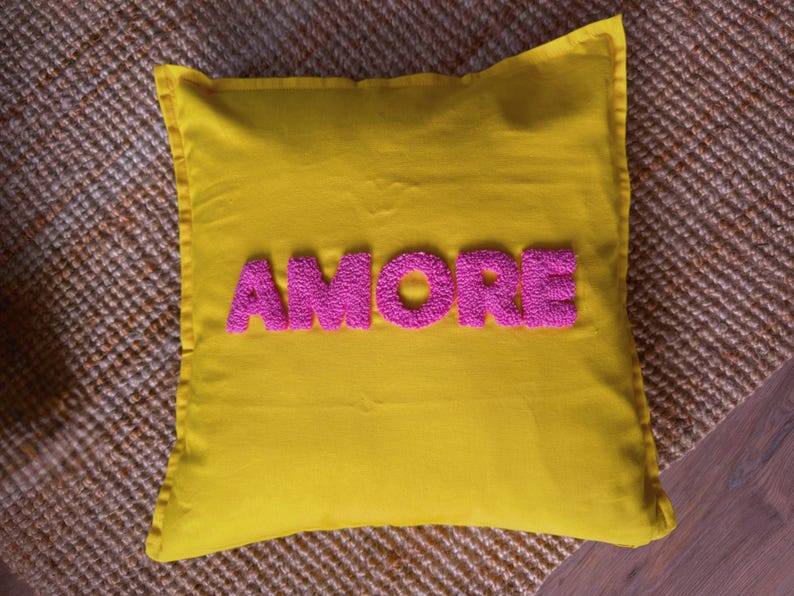 May include: A square yellow pillow with the word "AMORE" embroidered in pink, placed on a textured brown surface. The pillow has a simple design, with a solid yellow background and the word "AMORE" in a bold, textured font.