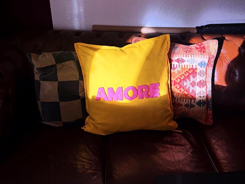 May include: Three decorative throw pillows on a brown leather couch. The center pillow is yellow with the word "AMORE" in pink. Other pillows feature a checkered pattern and a geometric design.