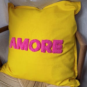 May include: A yellow decorative pillow with the word "AMORE" embroidered in bright pink, placed on a wooden chair with a woven seat. The pillow is square-shaped and adds a pop of color.