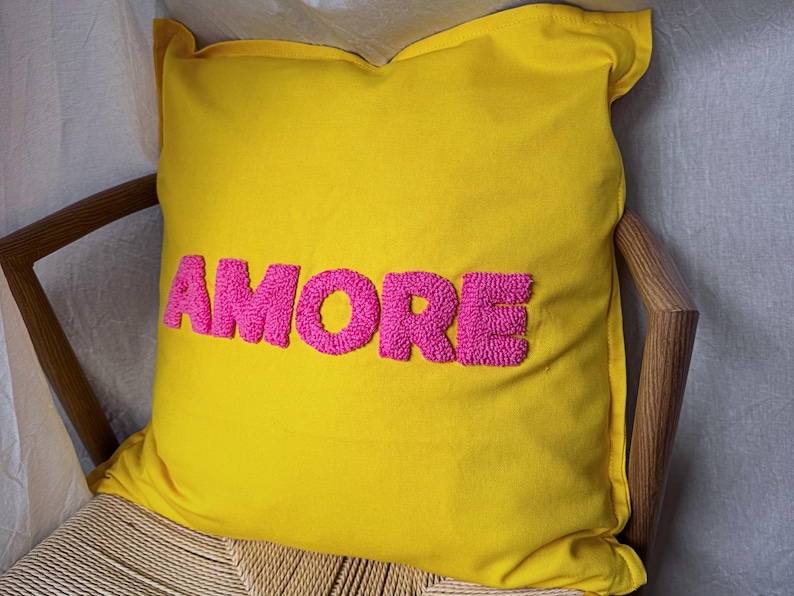 May include: A yellow decorative pillow with the word "AMORE" embroidered in bright pink, placed on a wooden chair with a woven seat. The pillow is square-shaped and adds a pop of color.