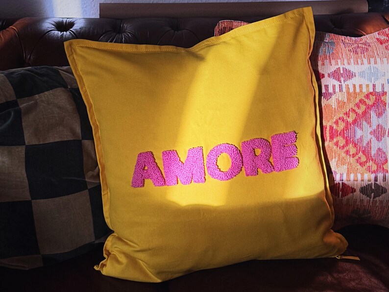 Könnte beinhalten: Ein senfgelbes Zierkissen mit dem Wort "AMORE" in Pink gestickt. Das quadratische Kissen liegt auf einem braunen Ledersofa, mit anderen gemusterten Kissen im Hintergrund.