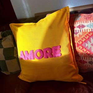 May include: A yellow decorative pillow with the word "AMORE" embroidered in pink. The pillow is rectangular and has a soft, plush appearance. Other pillows with different patterns are visible in the background.