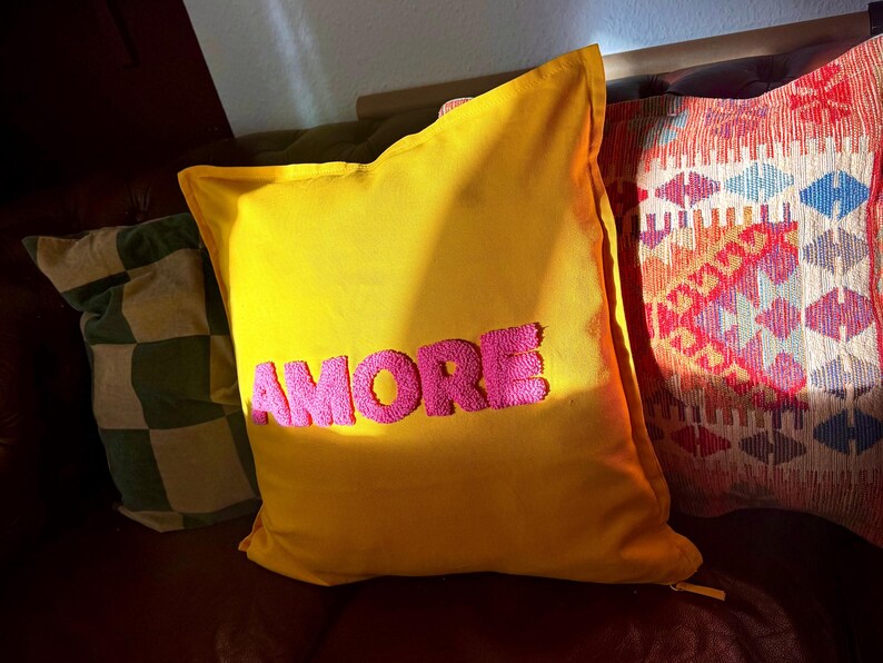 May include: A yellow decorative pillow with the word "AMORE" embroidered in pink. The pillow is rectangular and has a soft, plush appearance. Other pillows with different patterns are visible in the background.
