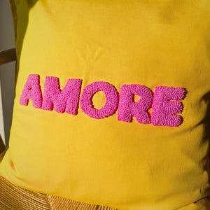 May include: Yellow decorative pillow with the word "AMORE" embroidered in bright pink, displayed on a wooden chair. The pillow's vibrant colors and textured lettering create a cheerful and inviting aesthetic.