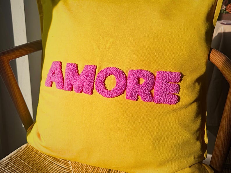 May include: Yellow decorative pillow with the word "AMORE" embroidered in bright pink, displayed on a wooden chair. The pillow's vibrant colors and textured lettering create a cheerful and inviting aesthetic.