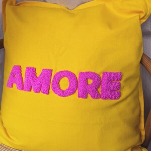 May include: A yellow decorative pillow with the word "AMORE" embroidered in bright pink, featuring a textured, raised design. The pillow is square and rests on a wooden chair.