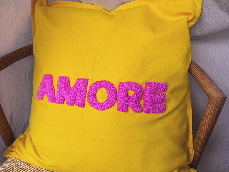 May include: A yellow decorative pillow with the word "AMORE" embroidered in bright pink, featuring a textured, raised design. The pillow is square and rests on a wooden chair.