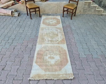 Op de afbeelding: Een lange, off-white loper met een vervaagd, verweerd patroon. Het tapijt heeft een subtiele, lichtroze kleur in het hele ontwerp. Het tapijt ligt op een grijs betegeld terras.