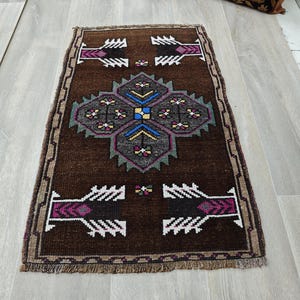 May include: A rectangular brown rug with a central geometric design in grey, blue, and purple. The rug features repeating patterns in white and purple on a dark brown background, with a fringed border. The rug is approximately 91 cm long and 61 cm wide.