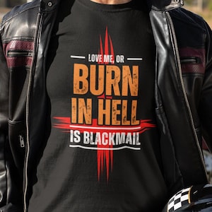 Puede incluir: Un hombre con una camiseta negra con el texto "LOVE ME, OR BURN IN HELL IS BLACKMAIL" en letras naranja y rojas. Lleva una chaqueta de cuero negra y pantalones vaqueros azules. Sostiene un casco de moto negro con una raya a cuadros.