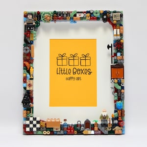 May include: A rectangular picture frame decorated with a variety of colorful plastic building bricks and miniature figures. The center features a yellow insert with the black text Little Boxes happy art and an illustration of three gift boxes.