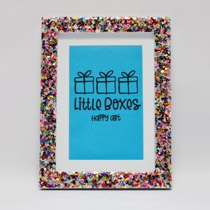 May include: A rectangular picture frame with a colorful glitter border. The frame holds a blue background with the text "Little Boxes happy art" and three gift box illustrations.