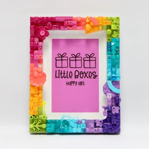 May include: A rectangular picture frame constructed from multicolored plastic building bricks in shades of pink, orange, yellow, green, blue, and purple. The center contains a bright pink insert with three black gift box icons and the text Little Boxes happy art.