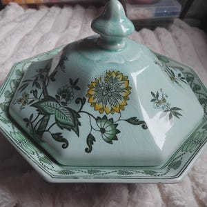 May include: A light blue ceramic serving dish with a lid. The dish has a hexagonal shape with a floral design in yellow, green, and black. The lid has a finial on top and the dish has handles on the sides.