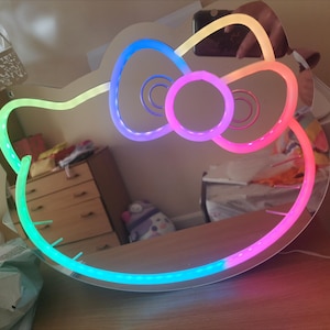 May include: A rainbow-colored LED light-up mirror shaped like Hello Kitty's face. The mirror is on a table and reflects the surrounding room.