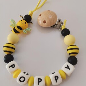 Op de afbeelding: Een fopspeenclip voor baby's met een bijen thema. De clip heeft een geel koord, een houten clip en een reeks kralen in geel, zwart en wit. De naam "POPPY" is gespeld met witte letterkralen en gele afstandhouders, en een bijenbedel.