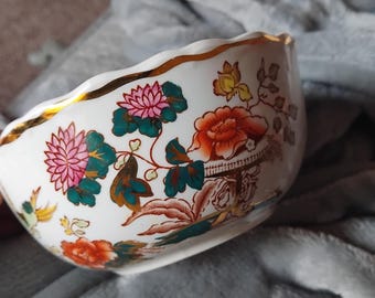 May include: White ceramic bowl with a scalloped gold rim. The bowl is decorated with colorful floral and foliage patterns in pink, orange, and teal. The bowl has a unique shape and is likely for decorative use.