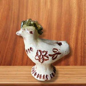 Può includere: Statuetta di uccello in ceramica con corpo color crema e motivi floreali marroni. L'uccello ha una cresta verde e poggia su una base a piedistallo. La statuetta è esposta su una superficie di legno.