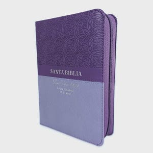 May include: A purple zippered Bible with a textured floral pattern on the upper cover. The cover features the text SANTA BIBLIA, Reina Valera 1960, Letra Grande, and 11 PUNTOS printed in white on a two-tone purple and light purple background.